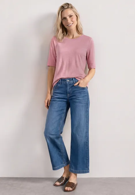Model draagt een roze t-shirt, blauwe wijde high-waist jeans en zwarte sandalen voor een neutrale achtergrond.