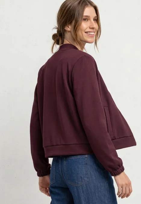 Jas met lange mouwen en een korter model rustic rouge Een glimlachende vrouw draagt een bordeaux jack en blauwe spijkerbroek en kijkt over haar schouder.