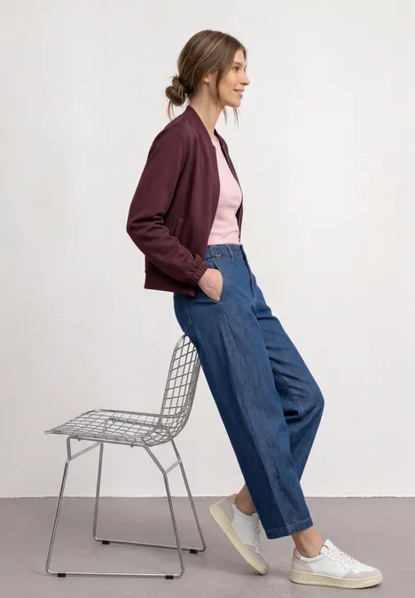 Jas met lange mouwen en een korter model rustic rouge Jonge vrouw leunt zijwaarts tegen een metalen draadstoel, draagt bordeaux jack, roze top, wijde jeans en witte sneakers.