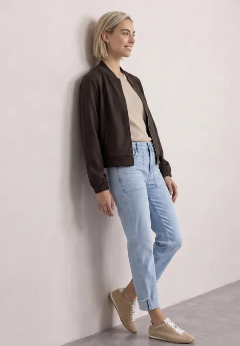 Jas met lange mouwen en een korter model black coffee Jonge vrouw met zwarte jas, beige shirt en blauwe jeans leunt ontspannen tegen een lichte muur.