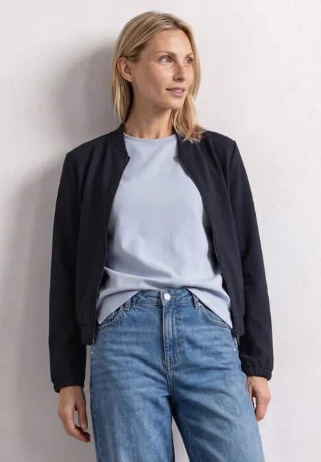 Jas met lange mouwen en een korter model darkest blue Vrouw in spijkerbroek, lichtblauw shirt en zwarte jas leunt ontspannen tegen een witte muur.