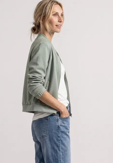 Jas met lange mouwen en een korter model eucalyptus green Vrouw in een groen bomberjack, wit shirt en blauwe jeans, staand in zijaanzicht met een lichte glimlach.
