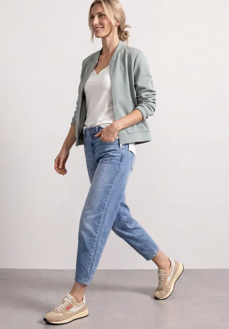 Jas met lange mouwen en een korter model eucalyptus green Vrouw in een grijs bomberjack, wit T-shirt, blauwe jeans en beige sneakers die ontspannen loopt.
