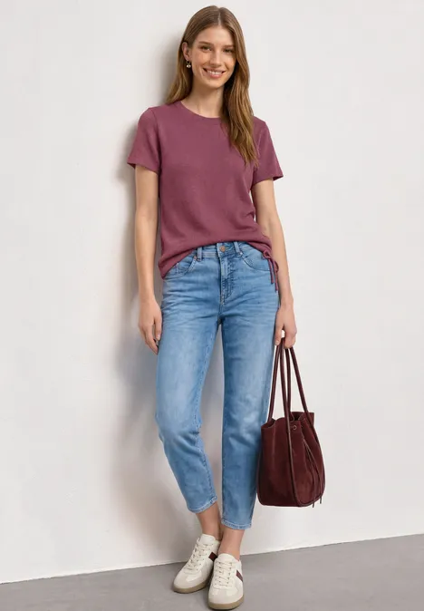 Vrouw leunt tegen witte muur, draagt bordeauxrood T-shirt, lichtblauwe jeans, witte sneakers en bijpassende tas.