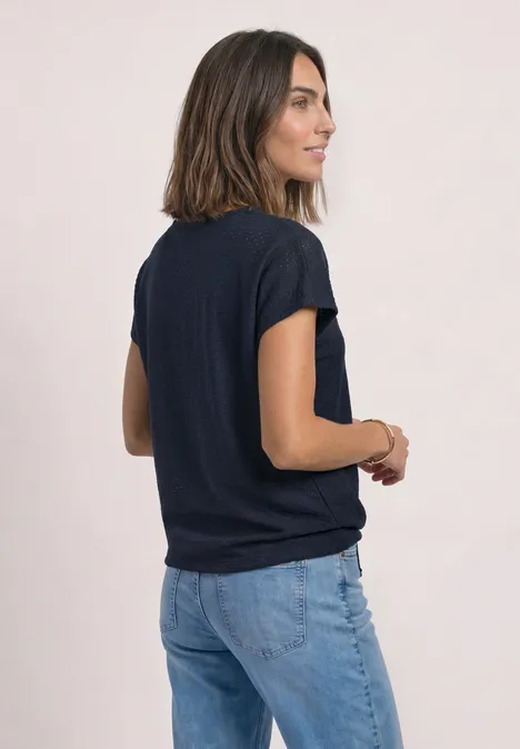 Kobieta stoi bokiem, ma na sobie granatowy T-shirt i dżinsy na jasnym tle.