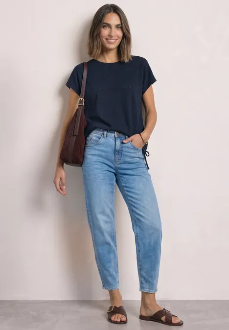 Uśmiechnięta kobieta na jasnym tle, w granatowym t-shircie, dżinsach, brązowych sandałach i z torebką na ramieniu.