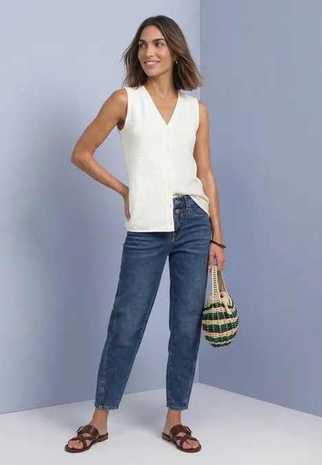Top mit V-Neck und dekorativer Knopfleiste off white Frau in weißer ärmelloser Bluse, Jeans und Sandalen mit Korbtasche vor lila Hintergrund.