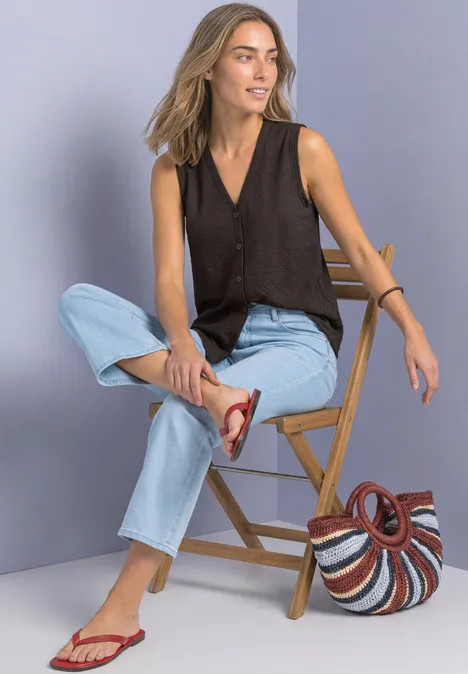 Frau in schwarzem Top, hellen Jeans und roten Sandalen sitzt lässig auf einem Holzstuhl, Korbtasche daneben.