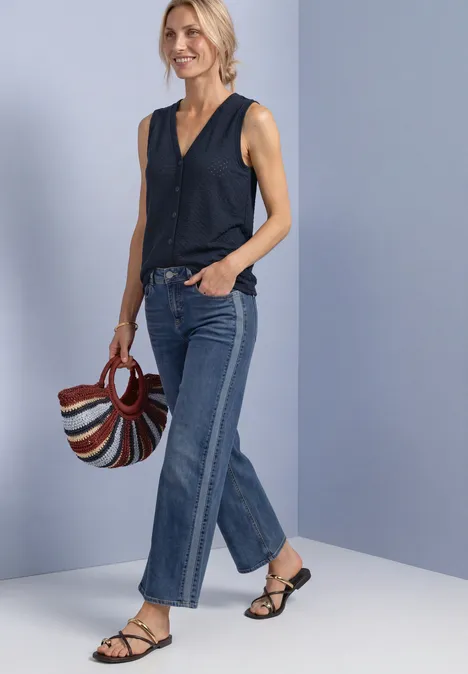 Haut à col en V et patte de boutonnage décorative darkest blue Frau trägt ärmellose schwarze Bluse, weite Jeans, Sandalen und hält eine bunte Tasche vor neutralem Hintergrund.