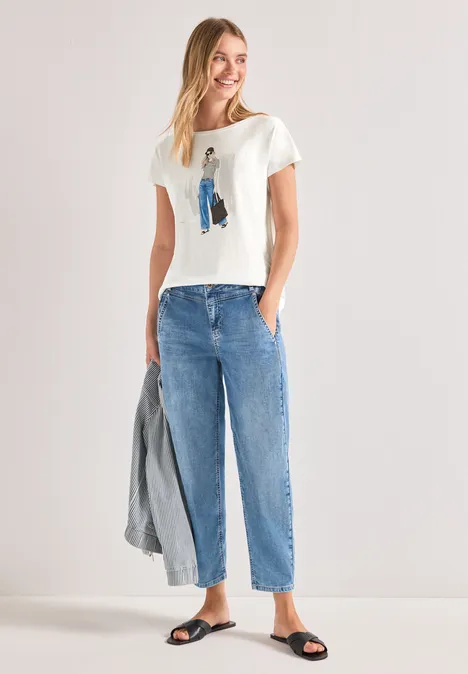 Frau in weißem T-Shirt, lockeren blauen Jeans und Sandalen hält eine graue Jacke.