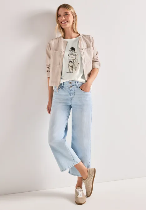 Vrouw in lichte jeans, wit grafisch t-shirt, beige blazer en loafers, staat nonchalant.