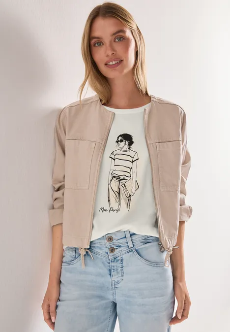 Vrouw in blauwe jeans, wit t-shirt met schetsprint en beige bomberjack voor witte achtergrond.