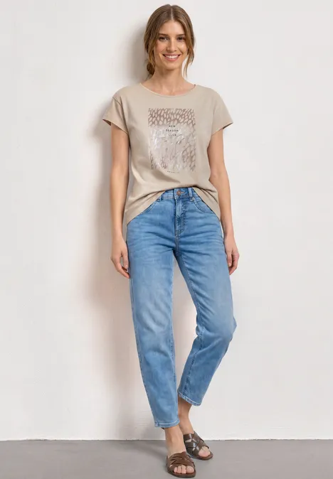 T-Shirt mit Rundhals und Foliendetails clay sand Frau in beigem T-Shirt mit Knoten, hellblauen Jeans und Sandalen vor weißer Wand.