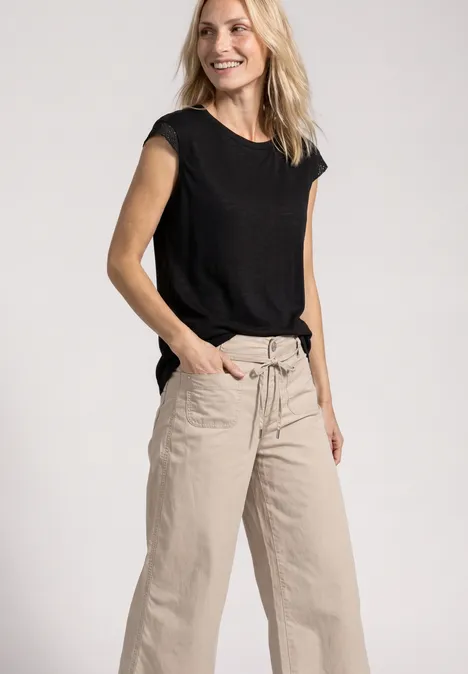 Een vrouw draagt een zwart T-shirt en beige wijde broek, lacht, handen in de zakken.