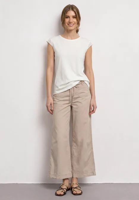 Jonge vrouw met wit t-shirt, beige wijde broek en sandalen, staand voor een neutrale achtergrond.