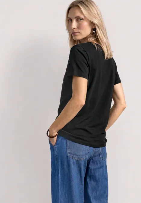 Vrouw in zwart T-shirt en blauwe jeans, rugzicht, één hand in de zak.