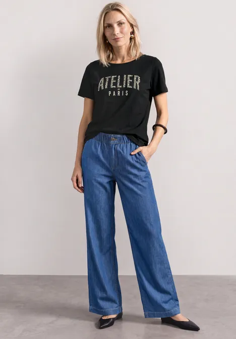 Vrouw met zwarte 'ATELIER PARIS' T-shirt, wijde blauwe jeans en zwarte schoenen.