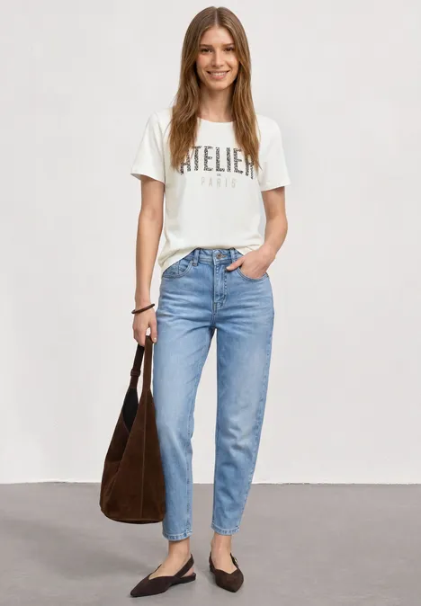 Frau in hellblauen Jeans und weißem 'Atelier'-T-Shirt, schwarze Ballerinas und braune Schultertasche.