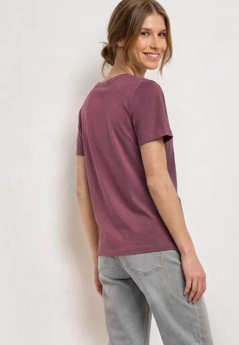 Shirt met ronde hals en opdruk op de voorkant dark clouded blush Vrouw in paars T-shirt en grijze spijkerbroek staat zijwaarts voor neutrale achtergrond