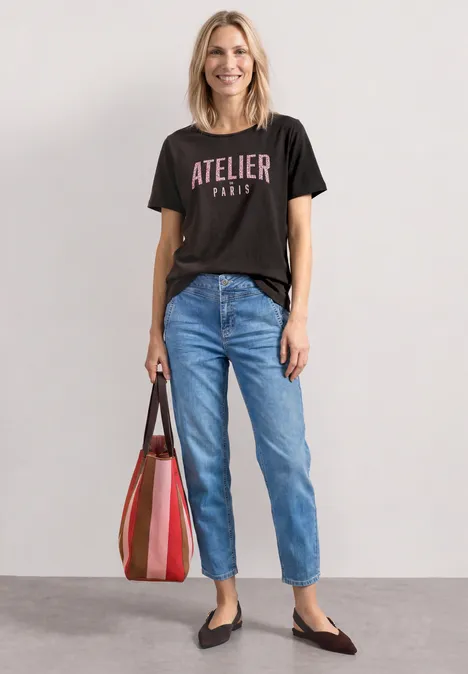 Femme porte un t-shirt noir «Atelier Paris», jean bleu, ballerines noires et grand cabas rayé.