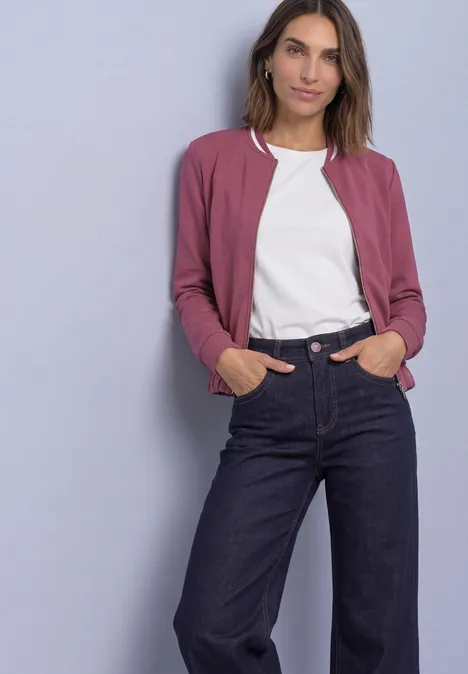 Eine Frau in einem weißen T-Shirt, rosa Cardigan und dunkelblauer, weiter Jeans vor hellem Hintergrund.