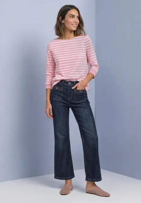 T-shirt à manches 3/4 et col bateau off white Frau mit rosa-weiß gestreiftem Pullover, weiten Jeans und Slippern vor neutralem Hintergrund.