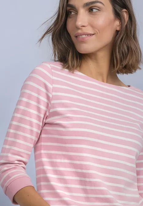 T-shirt à manches 3/4 et col bateau off white Frau mit schulterlangem Braunhaar trägt rosa-weiß gestreiftes Langarmshirt vor hellem Hintergrund.