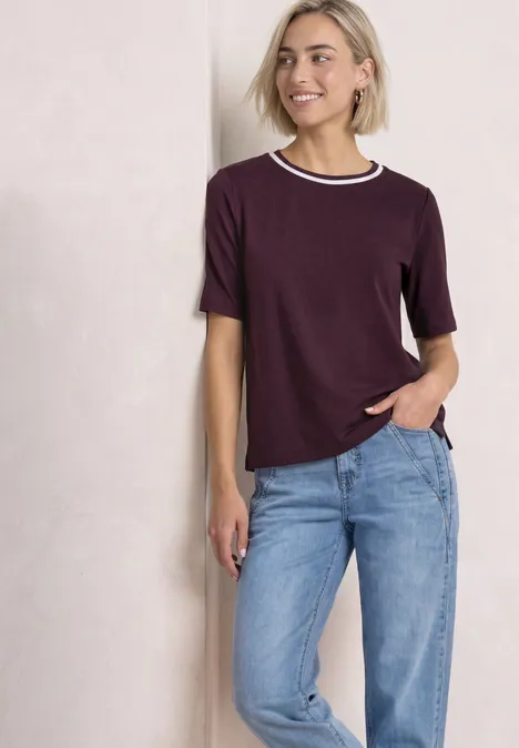 Uśmiechnięta kobieta w fioletowym t-shircie z białym kołnierzem i niebieskich jeansach opiera się o ścianę.