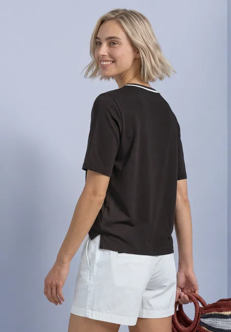 Młoda kobieta z blond boba, w czarnym t-shircie i białych szortach, spogląda przez ramię, czerwony dodatek