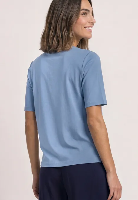 T-shirt à col rond avec détail contrasté northern blue Frau trägt hellblaues T-Shirt und dunkle Hose, lächelt seitlich vor hellem Hintergrund.