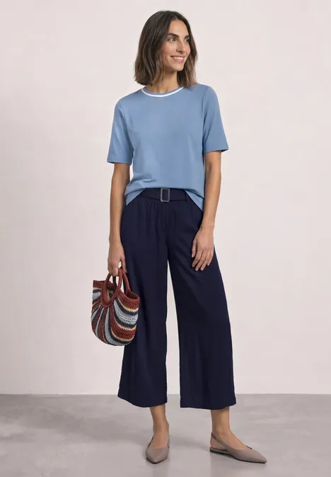 T-shirt à col rond avec détail contrasté northern blue Lächelnde Frau in blauem T-Shirt, weiter dunkelblauer Hose, trägt gestreifte Tasche und flache Schuhe.