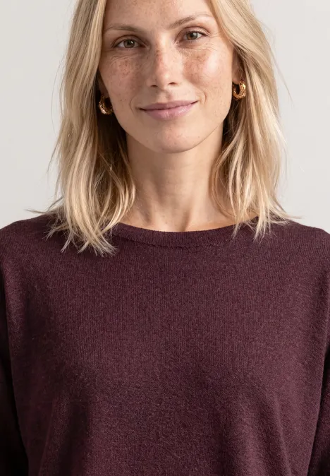 Eine blonde Frau mit leichtem Lächeln trägt einen dunkelroten Pullover vor hellem Hintergrund.