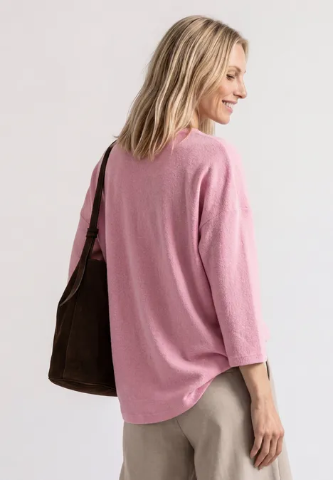 Eine Frau mit hellblondem Haar trägt ein rosafarbenes Oberteil und eine braune Schultertasche.