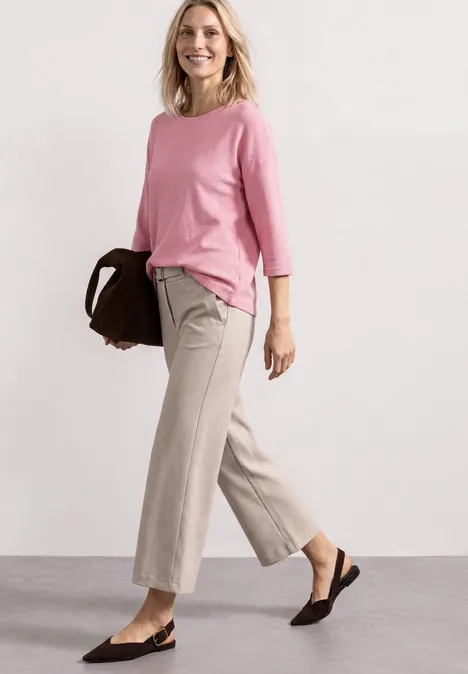 Lächelnde Frau in rosa Oberteil, beigen Culottes und schwarzen Ballerinas mit brauner Tasche.