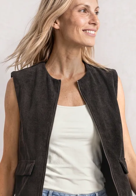 Vrouw met licht haar draagt een donkerbruine suède bodywarmer op een wit topje, lacht schuin.