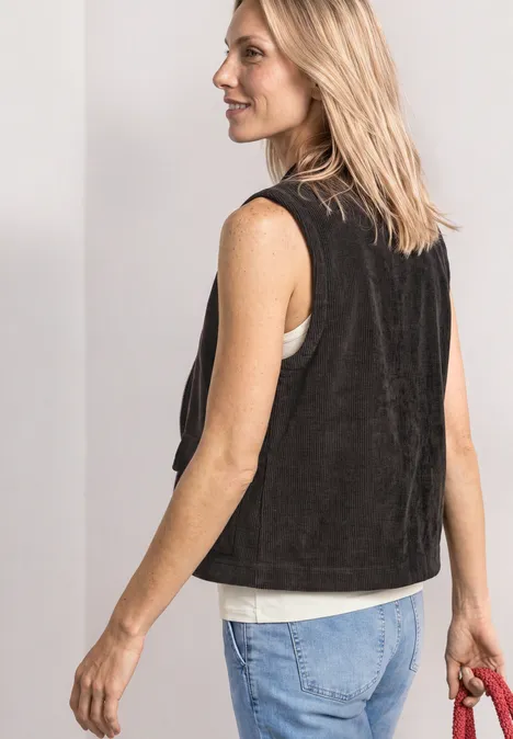 Vrouw in zijaanzicht met zwart vest, wit topje en jeans, houdt een rode tas vast.