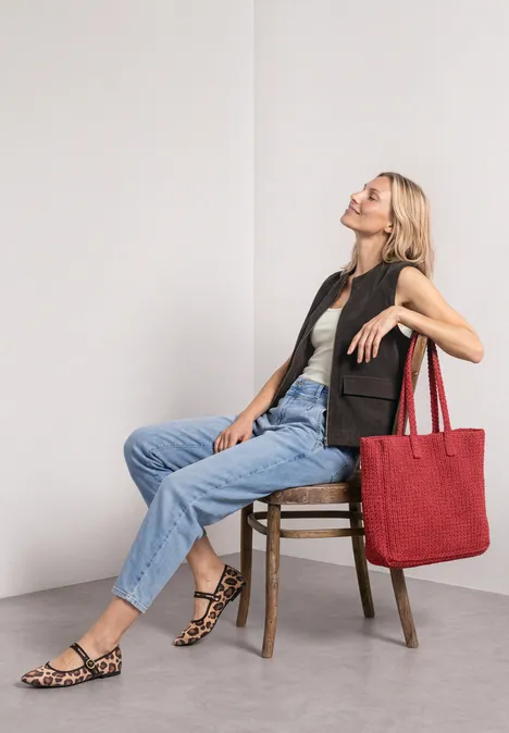 Vrouw zit ontspannen op een stoel voor een witte muur, draagt jeans, vest, sandalen en grote rode tas.