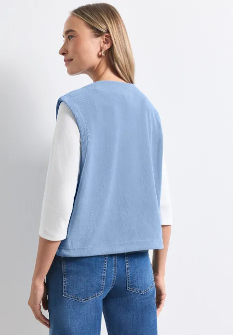 Gilet zippé à col rond effet velours côtelé northern blue Une femme de dos porte un gilet bleu clair sur un tee-shirt blanc et un jean devant un fond clair
