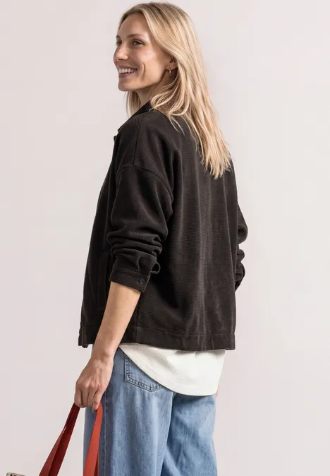 Jasje met ronde hals in corduroy-look black coffee Frau blickt lächelnd über die Schulter, trägt schwarze Jacke, weiße Bluse, blaue Jeans und rote Tasche.