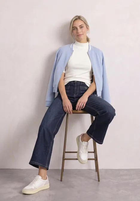 T-Shirt mit Turtle Neck in Rippstruktur off white Frau sitzt lässig auf Hocker, trägt weiße Sneaker, weite Jeans, weißen Rollkragenpullover und hellblauen Cardigan.