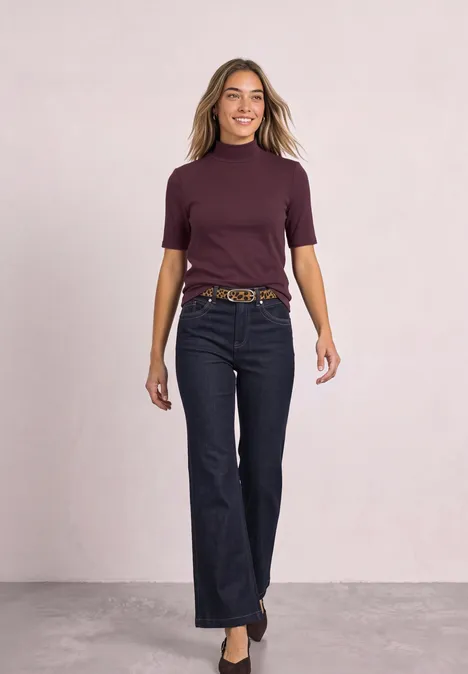 T-shirt à col roulé en maille côtelée rustic rouge Eine Frau im lila Kurzarmshirt und dunkler Jeans läuft selbstbewusst auf neutralem Hintergrund.