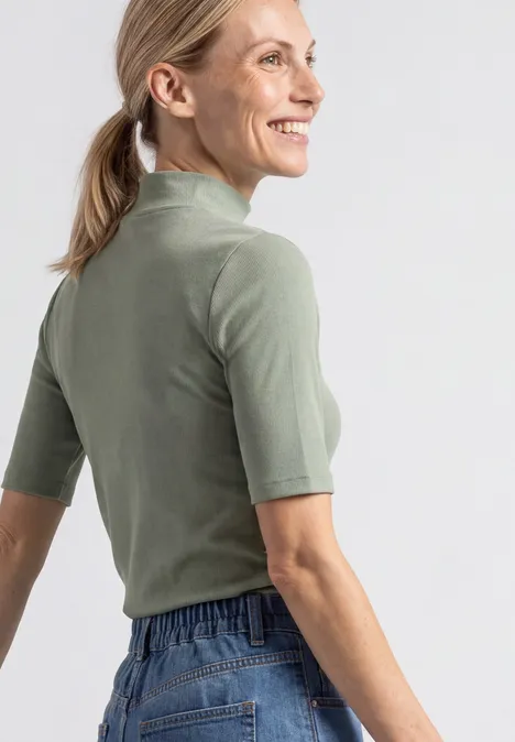 T-shirt à col roulé en maille côtelée eucalyptus green Eine Frau in einem olivgrünen Kurzarmshirt und Jeans schaut seitlich und lächelt.
