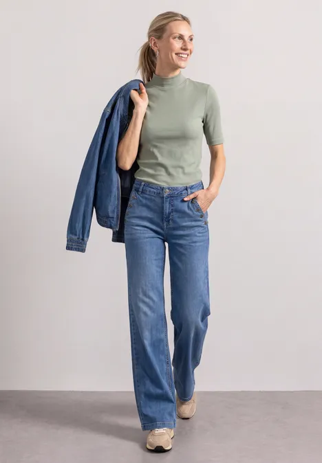 T-shirt à col roulé en maille côtelée eucalyptus green Eine Frau in grüner Kurzarmbluse und blauen Jeans, lächelnd und eine Jeansjacke über der Schulter tragend.