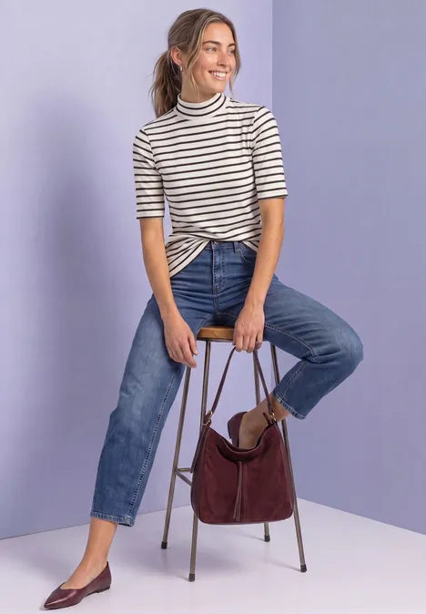 Jeune femme en marinière nouée et jean, assise sur un tabouret avec un sac bordeaux sur fond violet.
