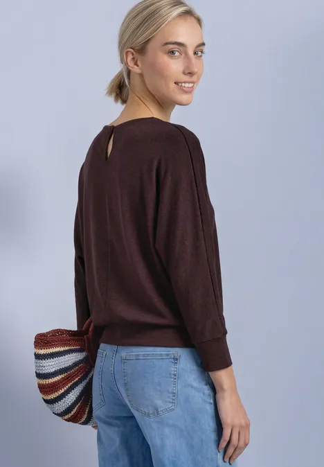 Frau in braunem Pullover und Jeans trägt eine bunte, gestreifte Tasche und lächelt über die Schulter.