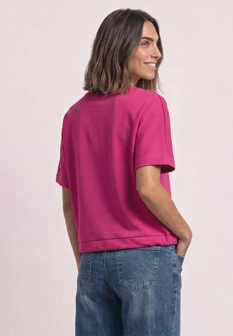 Lachende vrouw in zijaanzicht met een fuchsia t-shirt en spijkerbroek, handen in de zakken.
