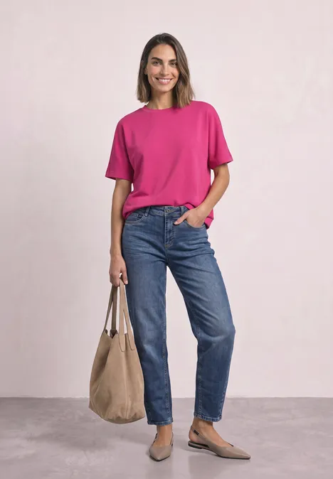 Glunderende vrouw in roze T-shirt en blauwe jeans met een beige tas voor een lichte achtergrond.