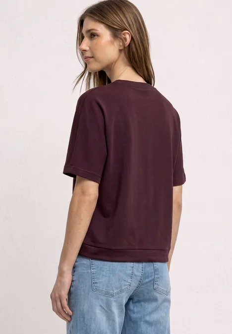 T-shirt à col rond avec un aspect soyeux rustic rouge Jeune femme vue de dos et de profil, portant un t-shirt bordeaux et un jean bleu clair sur fond clair.