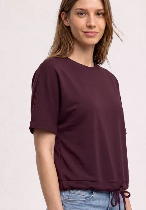 T-shirt à col rond avec un aspect soyeux rustic rouge Une jeune femme porte un T-shirt prune à manches courtes avec cordon de serrage et un jean bleu.