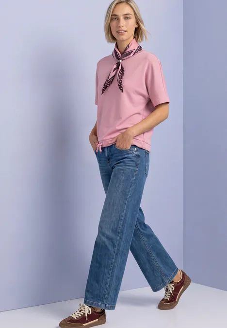 T-shirt à col rond aspect soyeux vintage blush Femme en haut rose, foulard, jean bleu et baskets devant un fond violet clair.